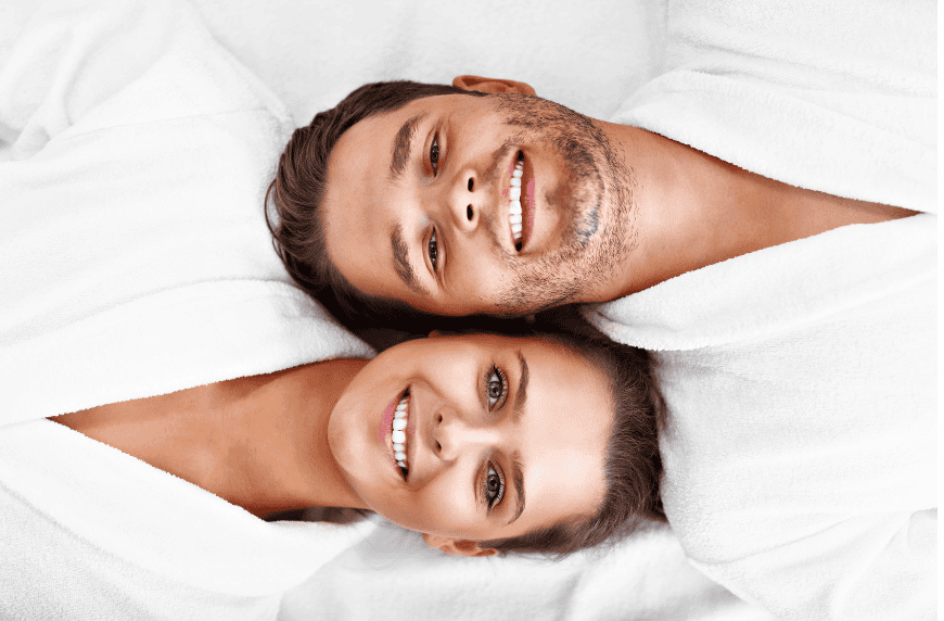 A smiling couple lying down in white robes, looking relaxed and happy.